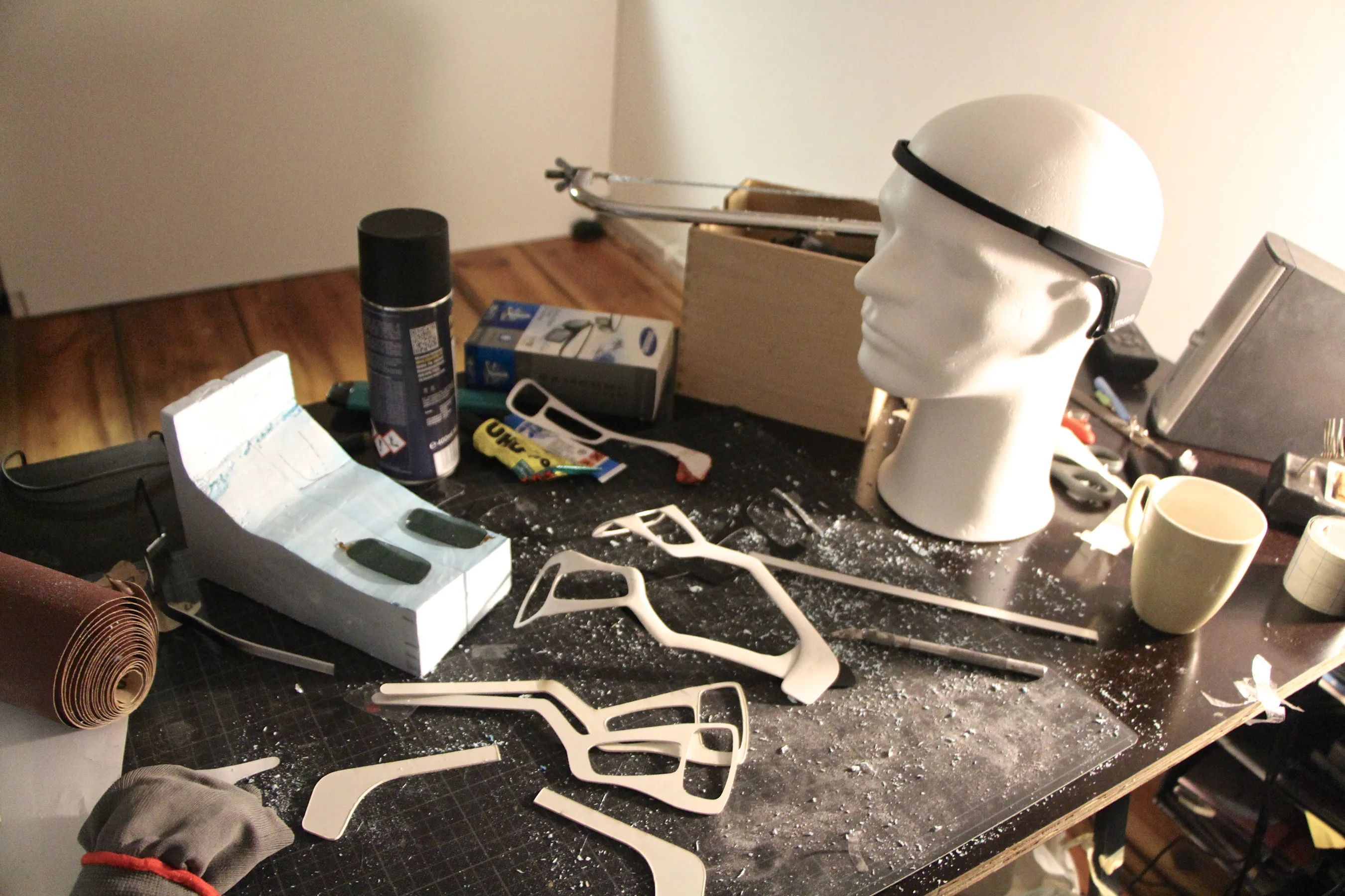 A cluttered workbench with thin white plastic cutouts of glasses frames, a white block holding two dark lenses, a spray can, a hacksaw and a mug, beside a white polystyrene head wearing a black strap