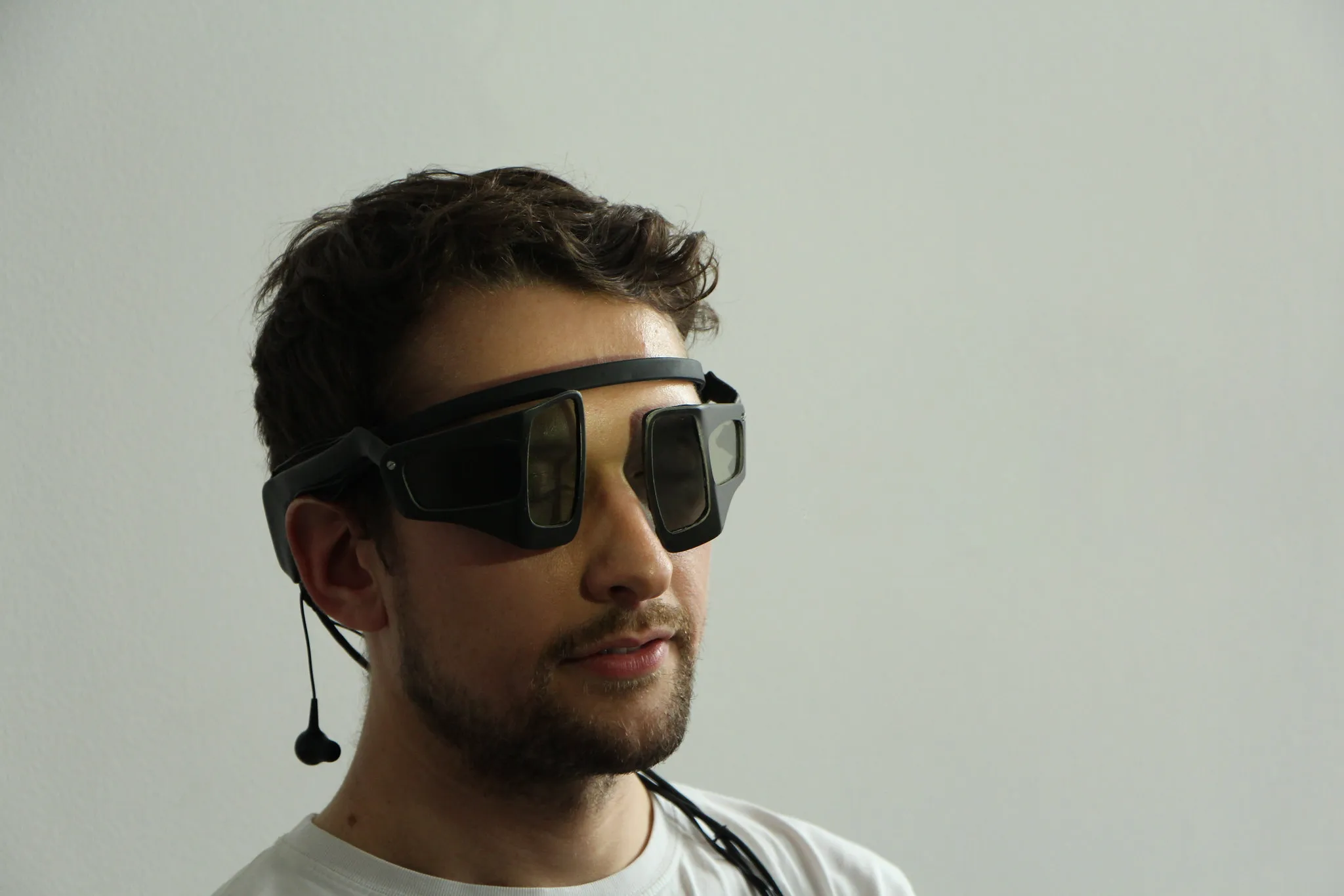 A young man wears the black liquid-crystal shutter glasses with their square lenses, an earphone hanging from a cable beside his ear, against a plain white wall
