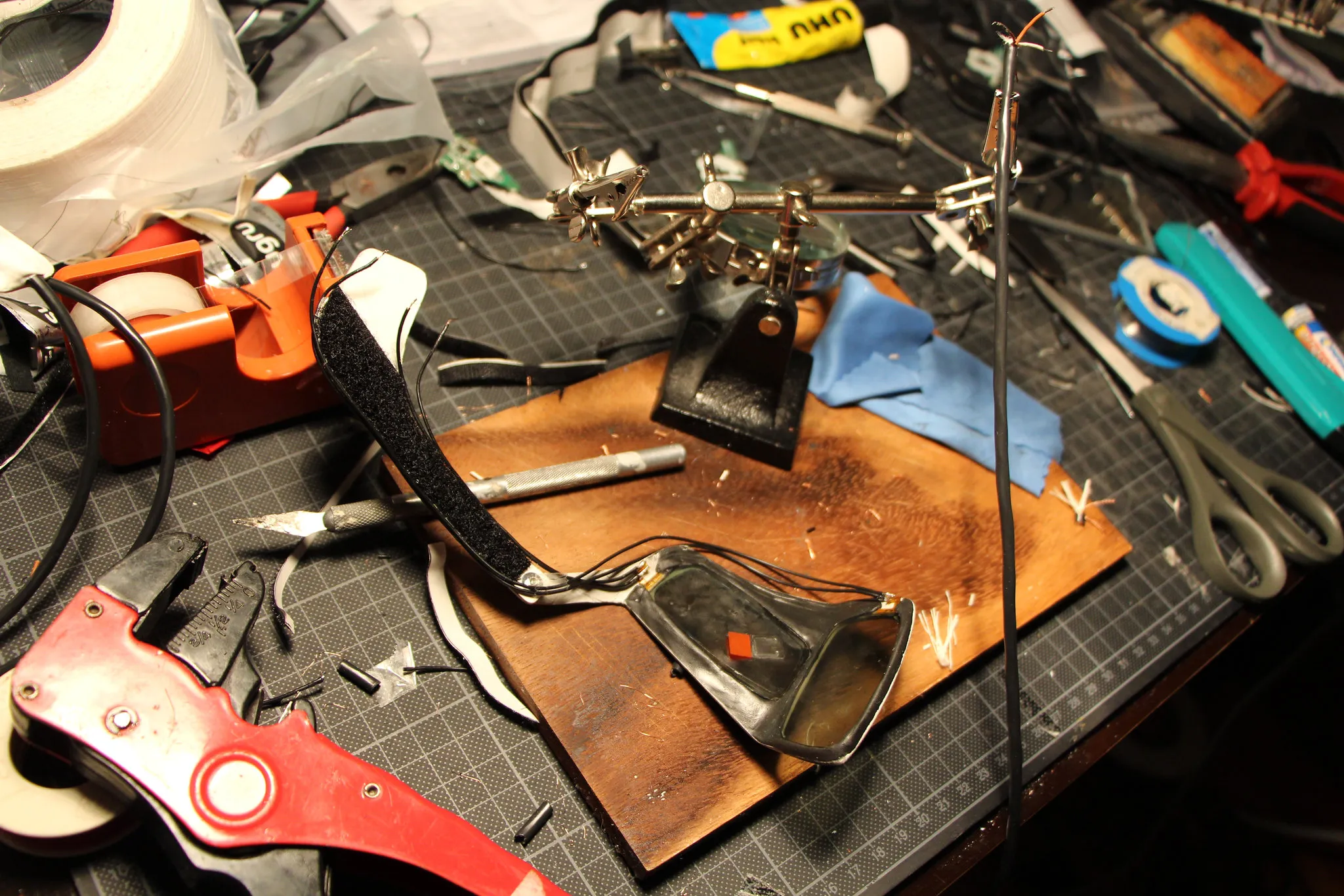 One half of the shutter glasses, its temple lined with Velcro, lies on a scorched wooden board beside a helping-hands clamp, thin wires soldered to its lens, surrounded by a wire stripper, tape, a scalpel and scissors