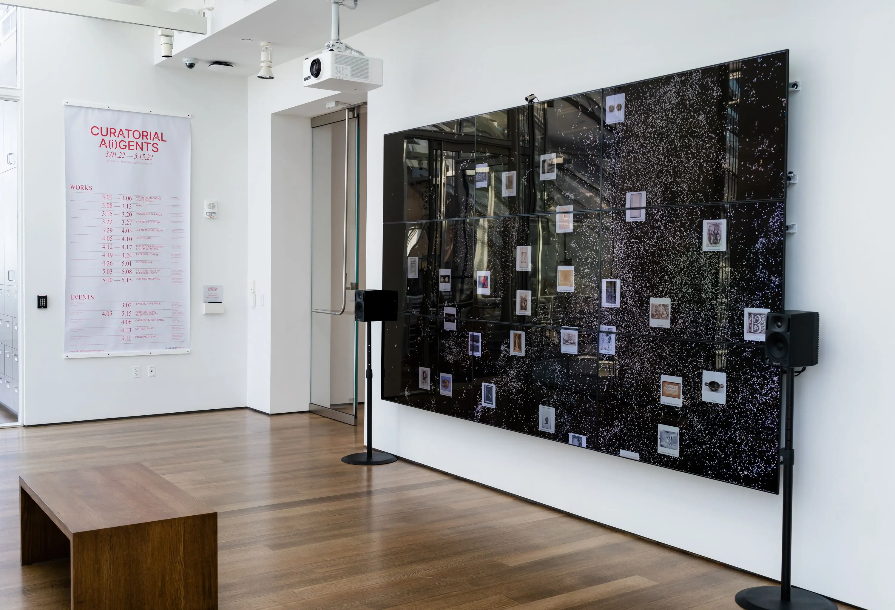 The Lightbox Gallery seen at an angle: the nine-monitor wall at right shows the collection as a field of white dots with object cards, between two speakers on stands; on the wall at left hangs the Curatorial A(i)gents program banner, listing Surprise Machines for 5.10–5.15, with a wooden bench in the foreground